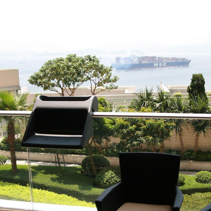 Ein Balkonblick mit einem schwarzen Stuhl und Halter, mit Blick auf üppiges Grün und ein Containerschiff auf schimmerndem Wasser.