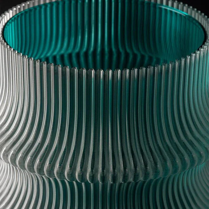 Nahaufnahme einer modernen Glasvase mit welliger, geriffelter Textur, die durchscheinende blaugrüne und klare Segmente vor einem dunklen Hintergrund aufweist.