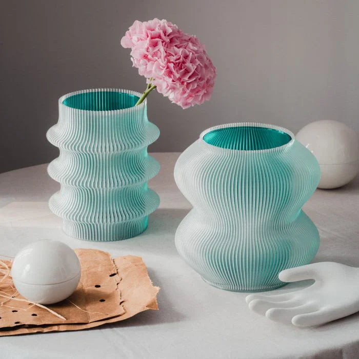 Zwei stilvolle Vasen mit strukturiertem mintgrünem Muster halten eine rosa Blume. Eine weiße Kugel und eine Handskulptur stehen in der Nähe auf einem neutralen Tisch.