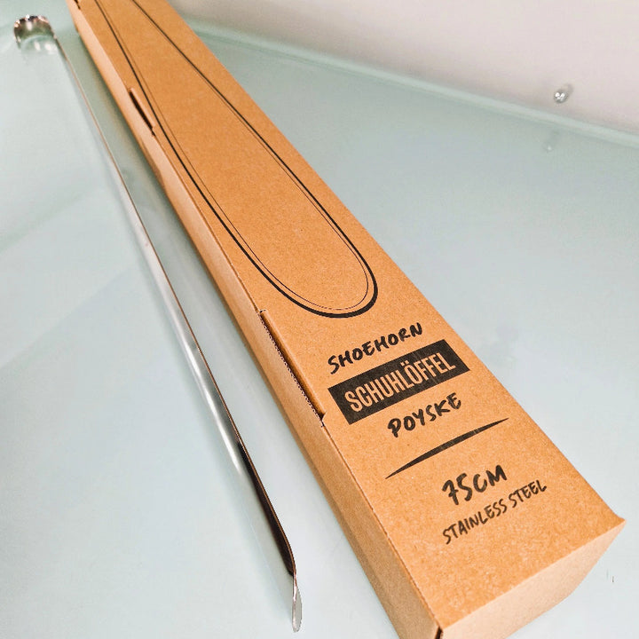 Schuhlöffel aus Edelstahl in einer einfachen braunen Box mit der Aufschrift „75 cm“ auf einer reflektierenden Oberfläche, konzipiert für einfaches Anziehen der Schuhe.
