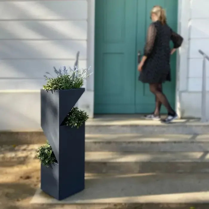 Ein moderner, geometrischer Blumentopf voller grüner Pflanzen und blauer Blumen steht auf Stufen neben einer Frau, die sich einer blaugrünen Tür nähert.