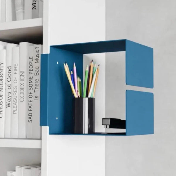 Auf einem modernen blauen Wandregal stehen ein schwarzer Stifthalter mit bunten Stiften und ein Hefter, neben gestapelten weißen Büchern.