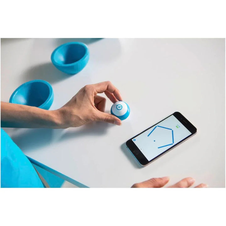 Hand hält kleinen programmierbaren Roboterball neben Smartphone mit Coding-App, blaue Schalen