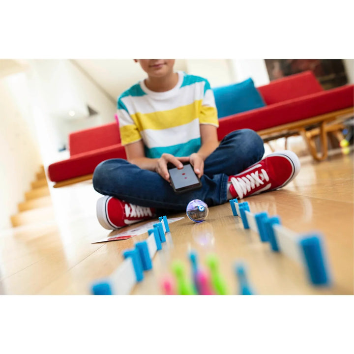 Kind steuert Sphero Roboterball mit Tablet im Wohnzimmer, bunte Dominosteine auf Holzboden