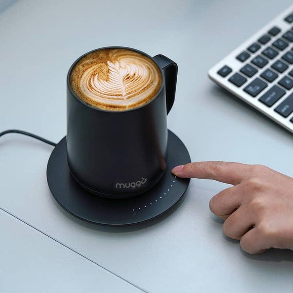 Eine Hand ist bereit, einen Untersetzer mit Temperaturregelung zu berühren, der eine schwarze Tasse mit Latte Art auf einem Schreibtisch neben einer Tastatur hält.
