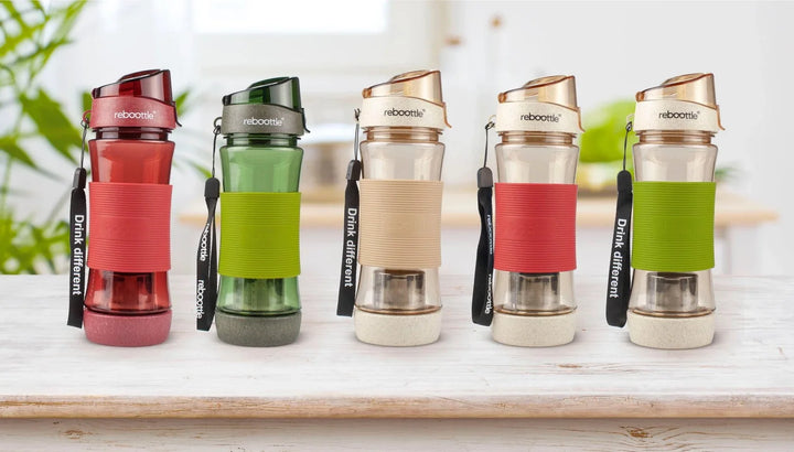 Fünf Reboottle Trinkflaschen aus Glas in verschiedenen Farben auf weißem Holztisch in heller Küche