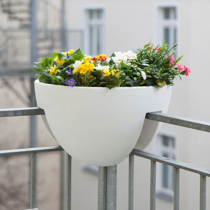 Ein weißer Balkonkasten voller leuchtender Blumen, sicher auf einem Metallgeländer eingebettet, vor einem verschwommenen städtischen Hintergrund.