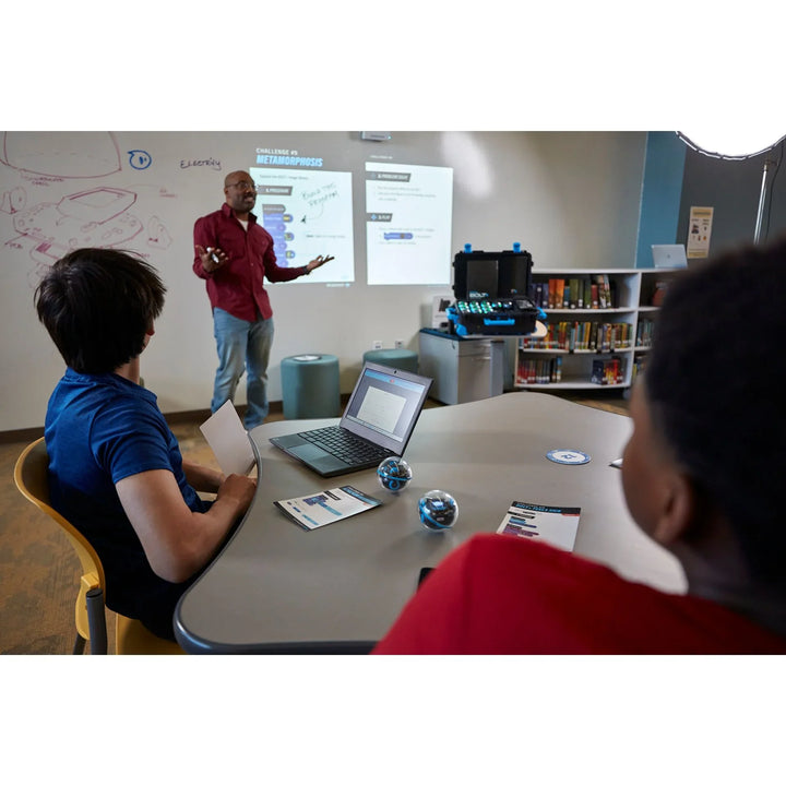 Lehrer erklärt Robotik-Programmierung im Klassenzimmer mit Laptops, Tablets und Sphero-Robotern.