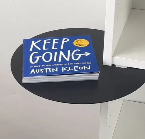 Buch 'Keep Going' von Austin Kleon auf schwarzem, runden Tisch neben weißem Regal
