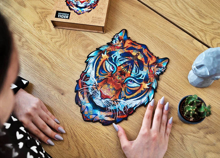 Auf einem Holztisch wird ein farbenfrohes, kunstvolles Tiger-Puzzleteil ausgestellt, das sanft von einer Hand berührt wird, und in der Nähe befinden sich Dekorationsgegenstände.
