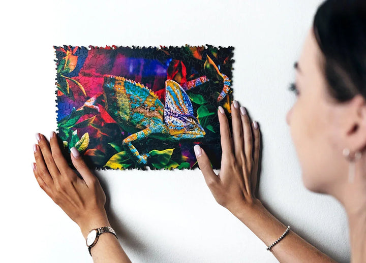 Eine Person hält ein lebendiges, farbenfrohes Chamäleon-Puzzle vor eine weiße Wand, umgeben von grünen Blättern und lebendigen Mustern.